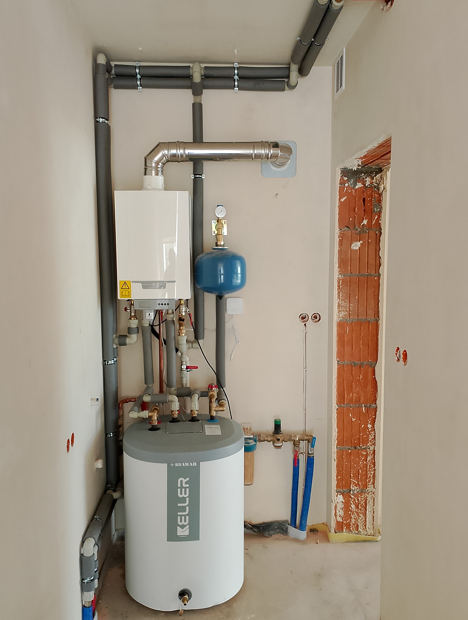 Biały piec gazowy dwufunkcyjny marki Keller, podłączony do instalacji CO z szarymi rurami izolowanymi termicznie, niebieski zbiornik wyrównawczy, widoczne elementy instalacji wodnej, ściana...