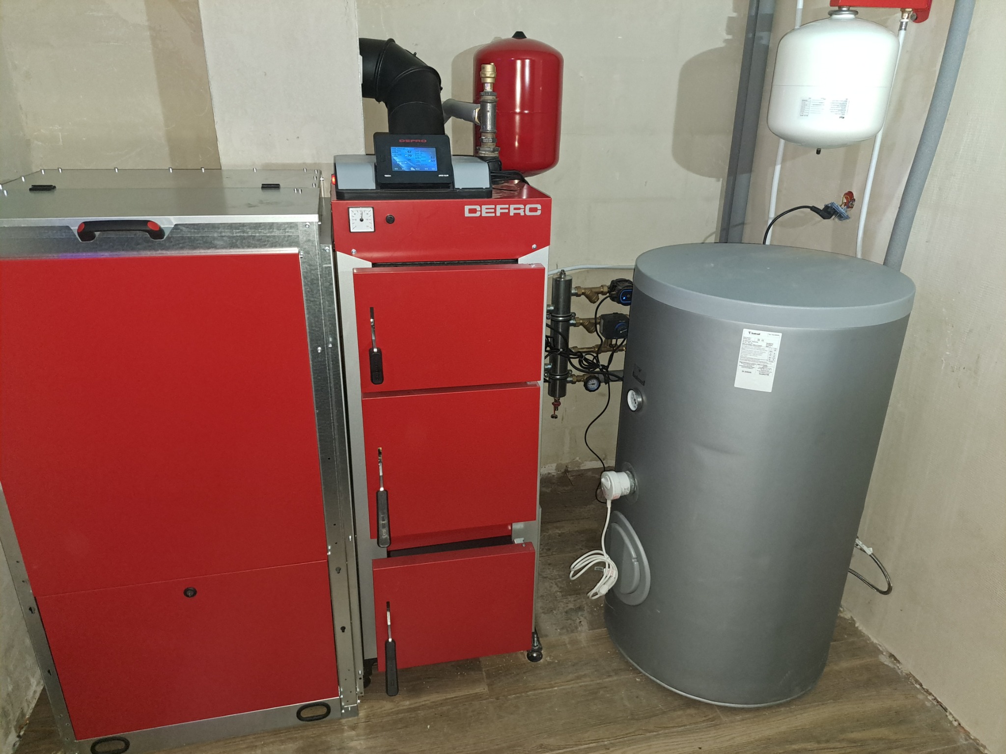 Czerwony piec CO marki Defro z zasobnikiem i zbiornikiem wyrównawczym w kotłowni, widoczne elementy instalacji.