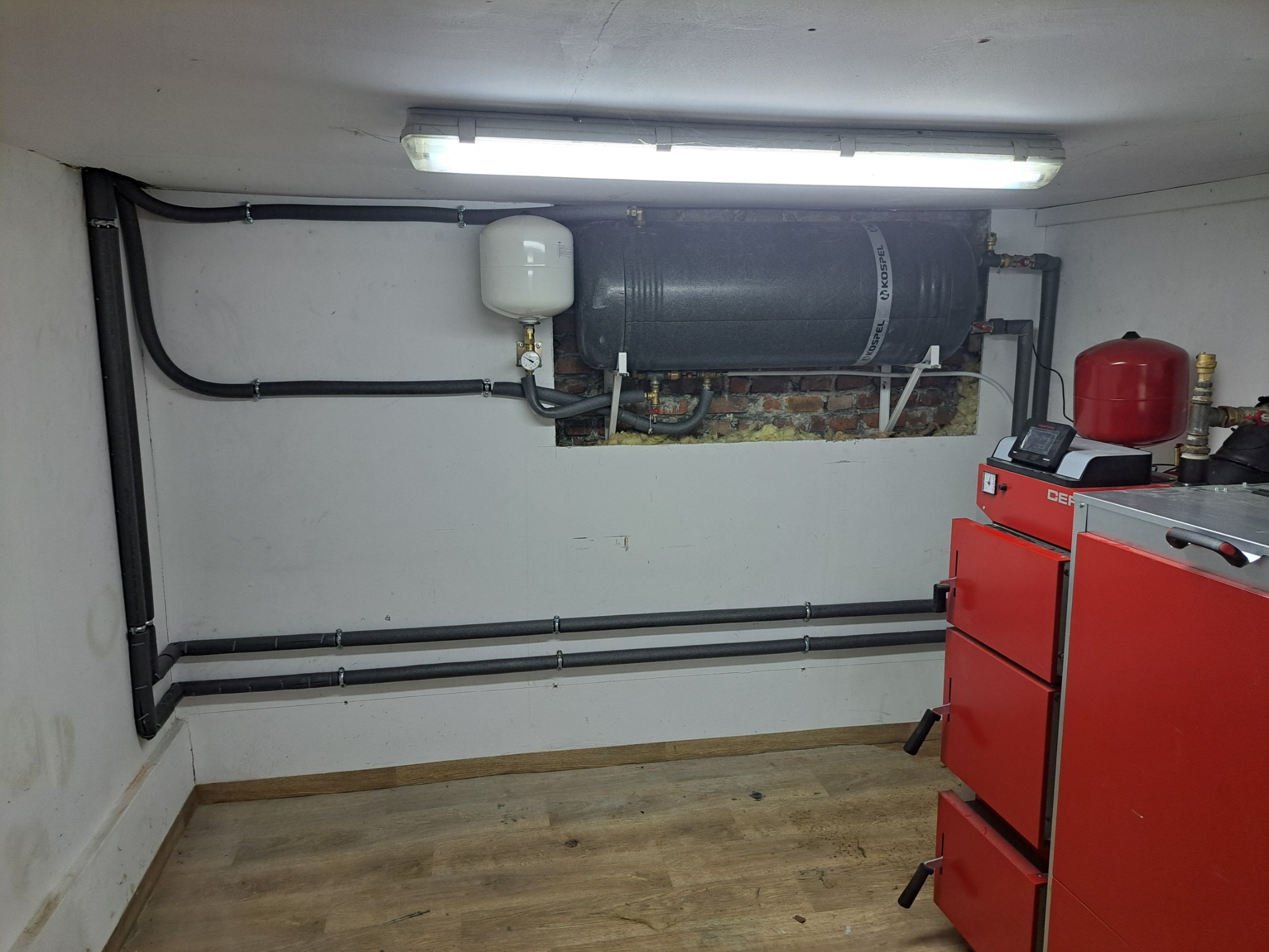 Kotłownia: czerwony piec CO z osprzętem, zbiorniki wyrównawcze i rury z izolacją na ścianie z ceglaną wnęką. Oświetlenie jarzeniowe, podłoga drewnopodobna.