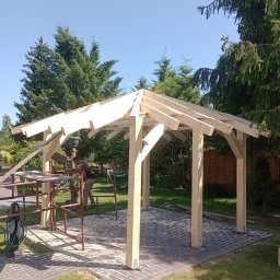 Dawid tomczak - Drewniana konstrukcja altany w trakcie budowy, widoczne rusztowanie i narzędzia. W tle zieleń drzew i błękitne niebo. Podłoże wyłożone kostką brukową.