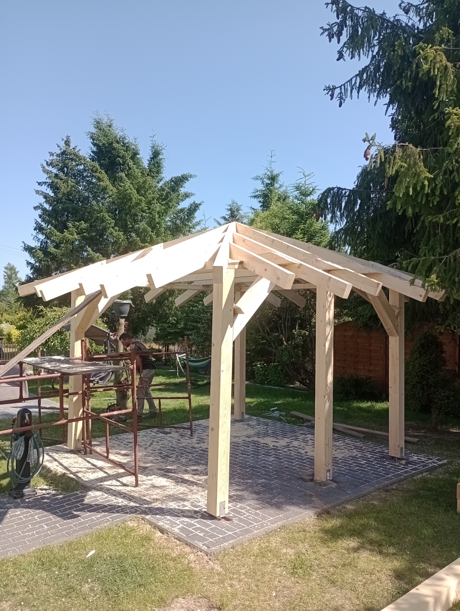 Drewniana konstrukcja altany w trakcie budowy, widoczne rusztowanie i narzędzia. W tle zieleń drzew i błękitne niebo. Podłoże wyłożone kostką brukową.