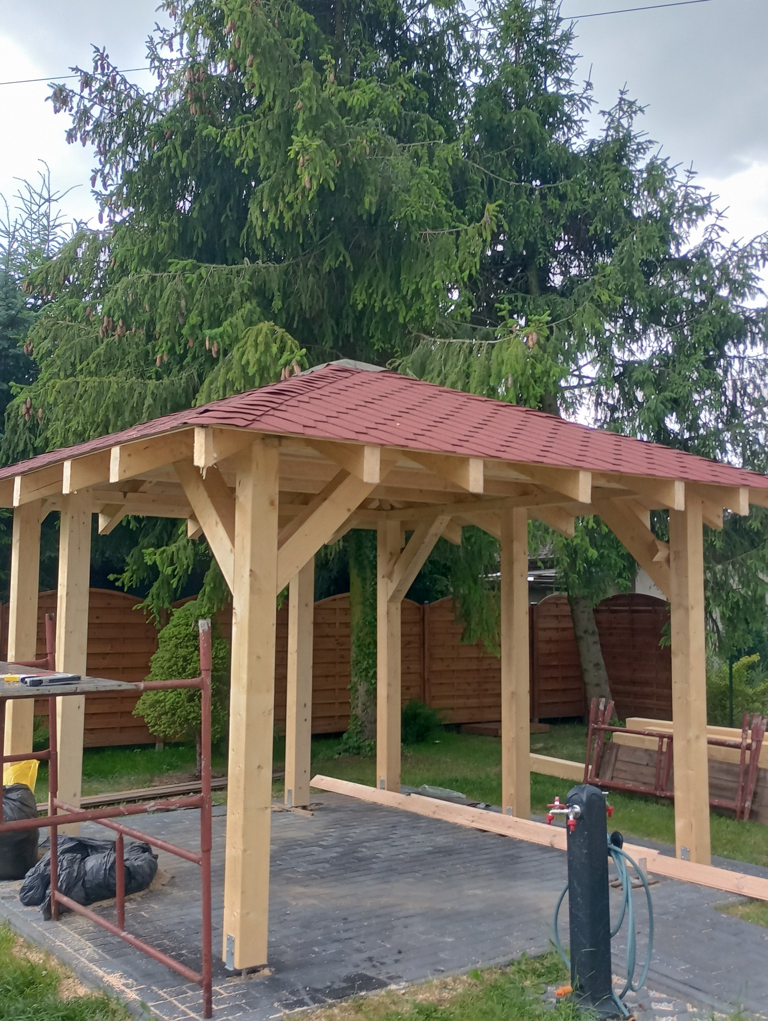 Drewniana altana z czerwonym dachem w trakcie budowy, widoczne rusztowania i narzędzia, tło stanowi zielony ogród z wysokimi drzewami i drewnianym ogrodzeniem.