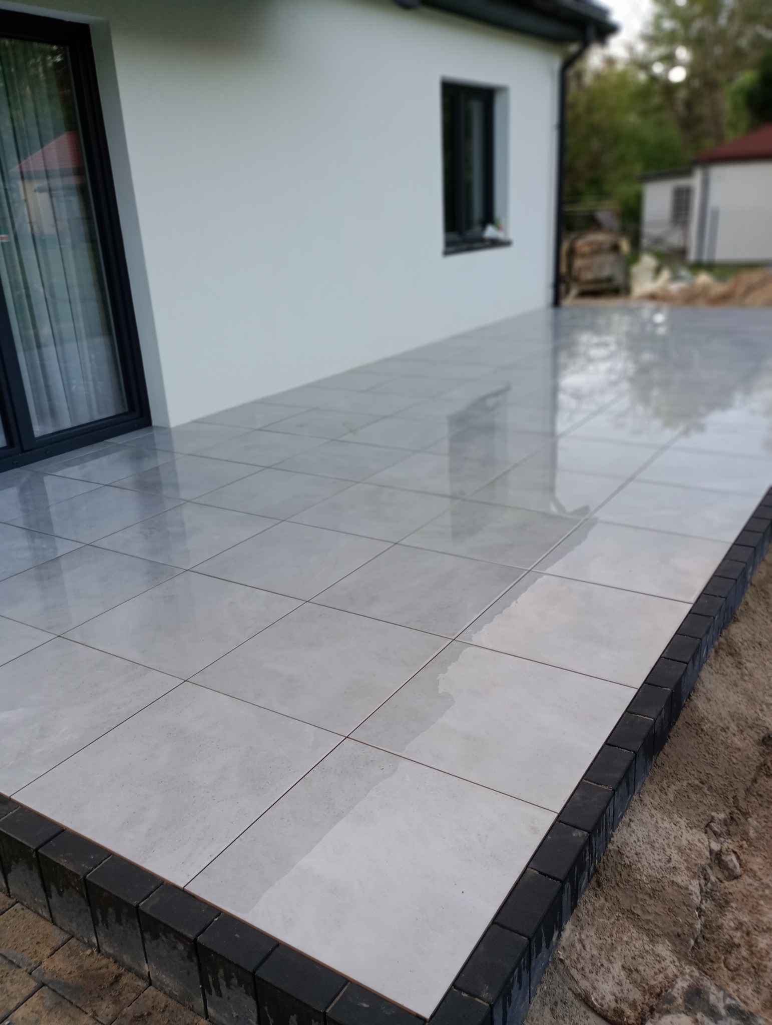 Jasnoszara, błyszcząca posadzka tarasowa z ciemną obwódką z kostki brukowej, przylegająca do białego budynku z czarną stolarką okienną i drzwiową.