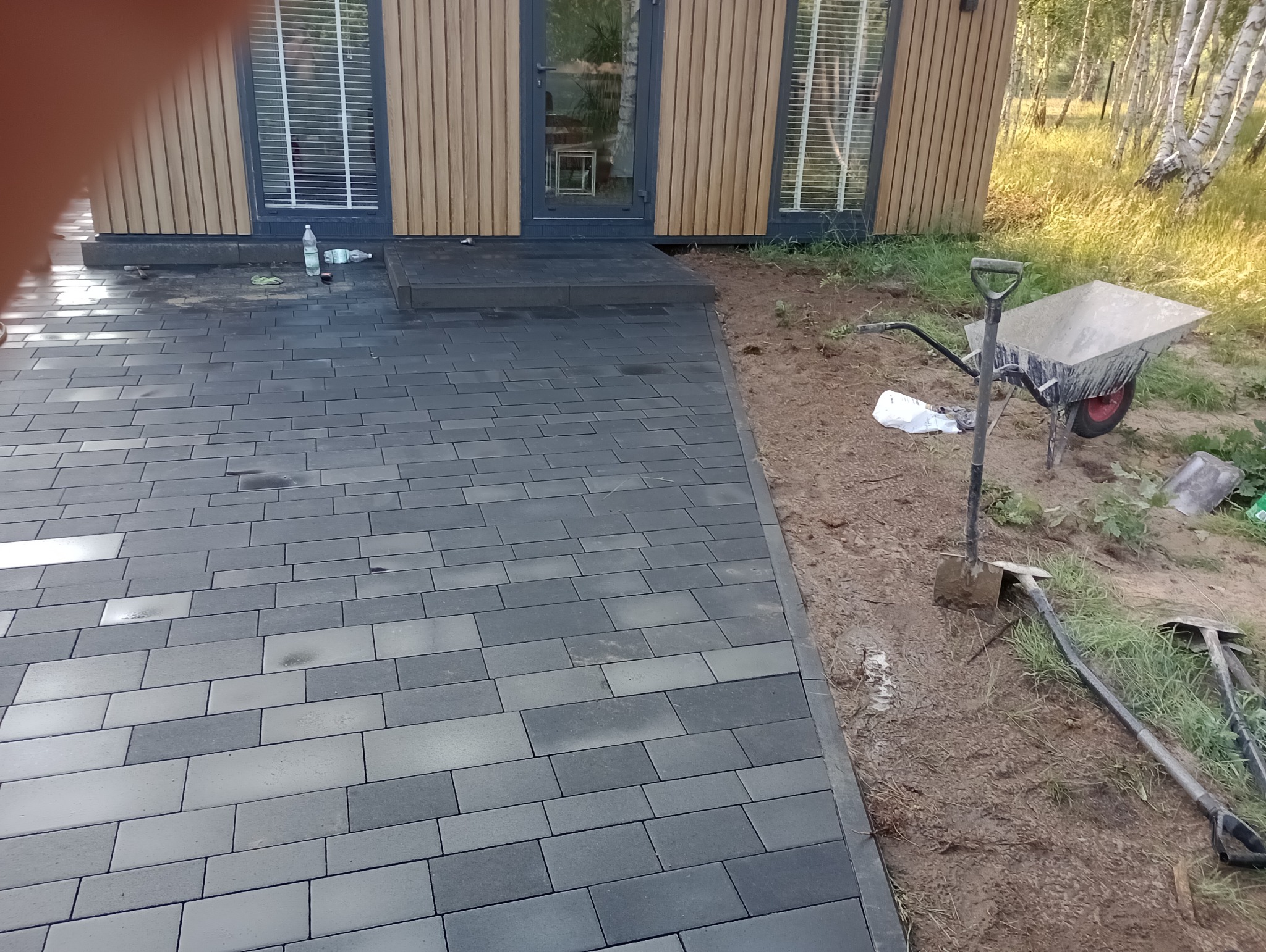 Ułożona, szara kostka brukowa przed budynkiem z drewnianą elewacją. Widoczne narzędzia brukarskie i taczka. Częściowo ukończona realizacja z bliska.