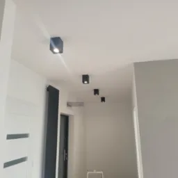 Nowoczesne oświetlenie sufitowe z czarnymi, kwadratowymi lampami punktowymi w białym korytarzu. Widoczny grzejnik panelowy w kolorze antracytowym i fragment drzwi.