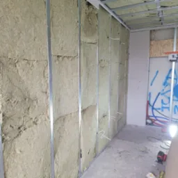 Ocieplenie ścian wełną mineralną w konstrukcji metalowej, widoczne przewody elektryczne wystające ze ściany, w tle fragment instalacji wodnej i płyty karton-gips
