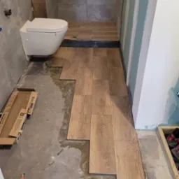 Układanie drewnopodobnych płytek podłogowych w trakcie remontu łazienki, widoczne kartony z płytkami, surowy beton i fragment podwieszanej toalety.