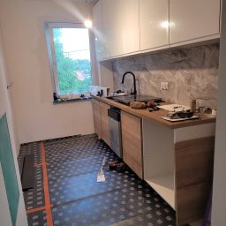 Montaż nowoczesnej kuchni: białe szafki wiszące, drewnopodobne blaty i fronty, czarna armatura. Na podłodze mata grzewcza. Widoczne narzędzia i akcesoria montażowe.