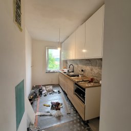 Wnętrze kuchni w trakcie montażu: białe szafki wiszące, drewnopodobne dolne, okno z widokiem na zieleń. Na podłodze narzędzia i materiały budowlane, ściany przygotowane do malowania.