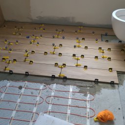 Układanie płytek imitujących drewno w łazience z zamontowaną toaletą. Widoczne żółte klipsy poziomujące i mata grzewcza pod płytkami.