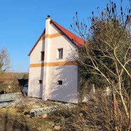 TLC KLUSSENBEDRIJF - Dwukondygnacyjny dom z beżową elewacją i pomarańczowym pasem, widziany z boku. Na pierwszym planie gałęzie drzewa i elementy rusztowania. Błękitne niebo w tle.