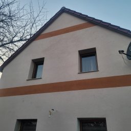 TLC KLUSSENBEDRIJF - Elewacja domu jednorodzinnego z dwoma pasami w kolorze brązowym. Widoczne okna i antena satelitarna. Na pierwszym planie gałęzie drzewa.