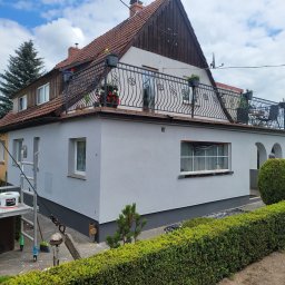 TLC KLUSSENBEDRIJF - Odnowiona elewacja domu z balkonem, świeżo po malowaniu na szaro. Widoczne rusztowanie, puszka farby Bolix i starannie przycięty żywopłot.