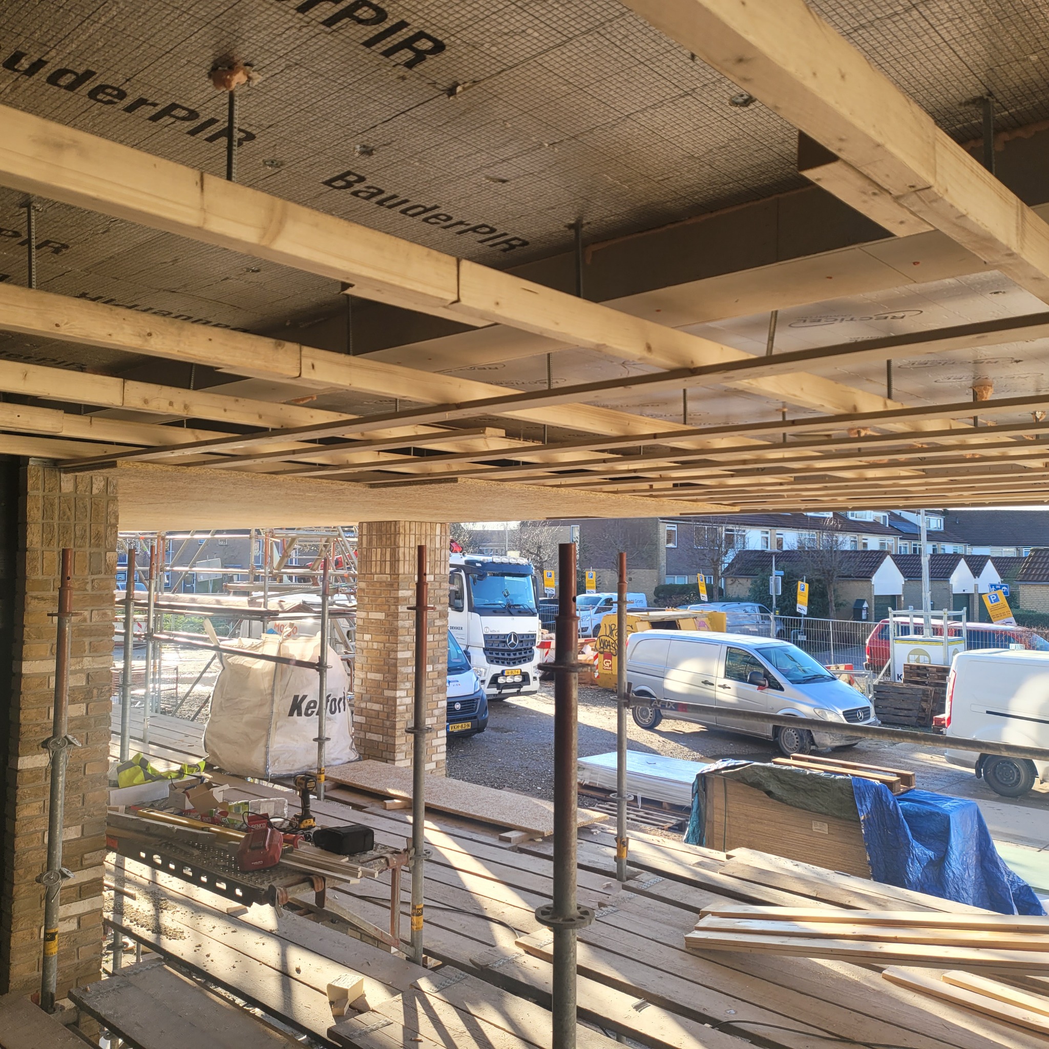 Montaż ocieplenia poddasza z widoczną konstrukcją drewnianą, materiałem izolacyjnym BauderPIR i elementami mocującymi, na tle placu budowy z rusztowaniami i zaparkowanymi pojazdami.
