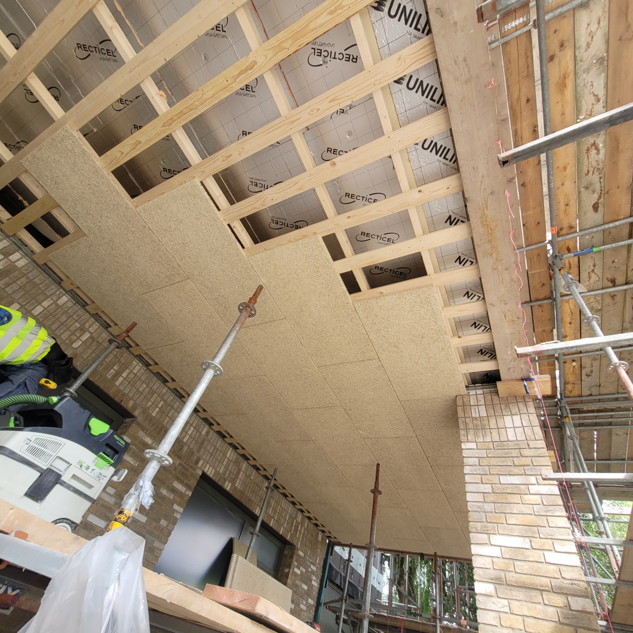 Montaż paneli izolacyjnych z włókna drzewnego na poddaszu, widoczne drewniane legary, folia izolacyjna z nadrukiem producenta, rusztowanie i narzędzia pracy.