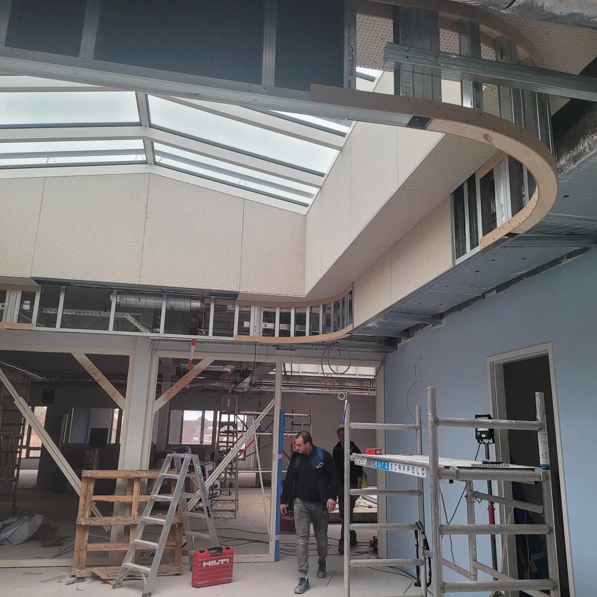 Montaż konstrukcji sufitu podwieszanego z płyt kartonowo-gipsowych, widoczne metalowe profile i drewniane elementy wykończeniowe, w tle okno dachowe i pracownik wchodzący do pomieszczenia.