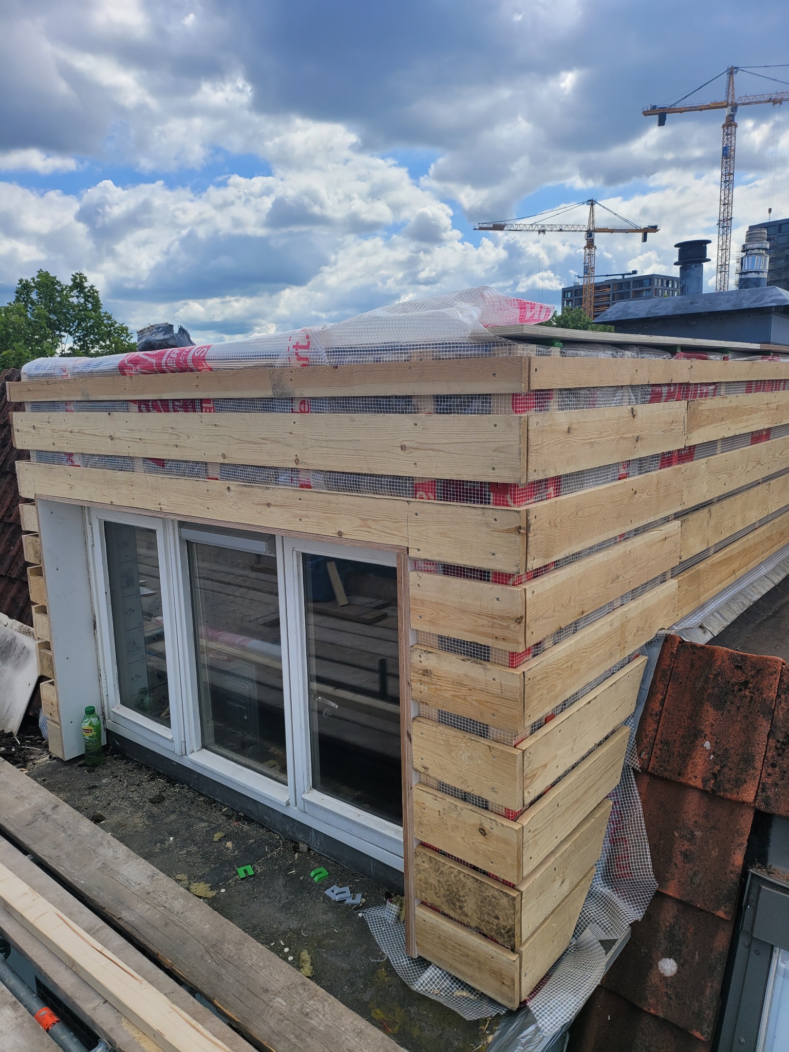 Drewniana konstrukcja szkieletowa obudowująca okno dachowe, zabezpieczona siatką i folią, widoczna na tle dachówek i nieba z chmurami oraz żurawi budowlanych w oddali.