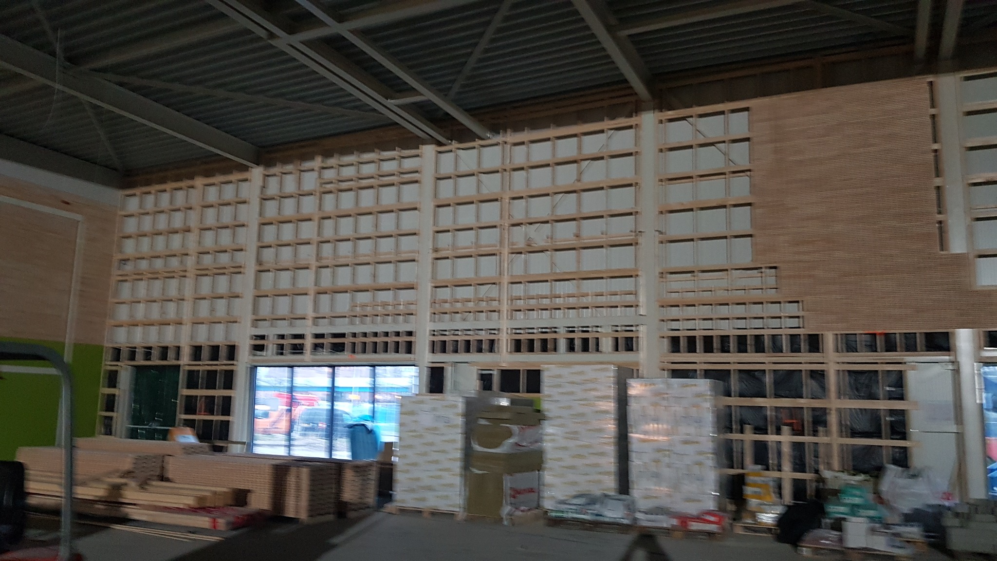 Wnętrze hali w trakcie prac wykończeniowych ścian: widoczna konstrukcja szkieletowa z drewna, częściowo wypełniona materiałem izolacyjnym, z fragmentem obłożonym płytą drewnopodobną, w tle okna...