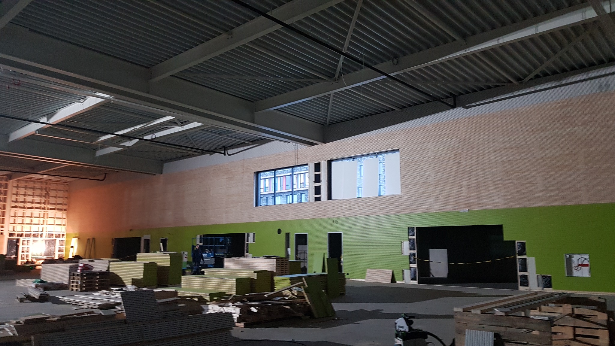 Wnętrze hali w trakcie prac wykończeniowych: drewniana boazeria kontrastuje z zielonymi ścianami, widoczne okna, materiały budowlane i narzędzia, surowy metalowy sufit.