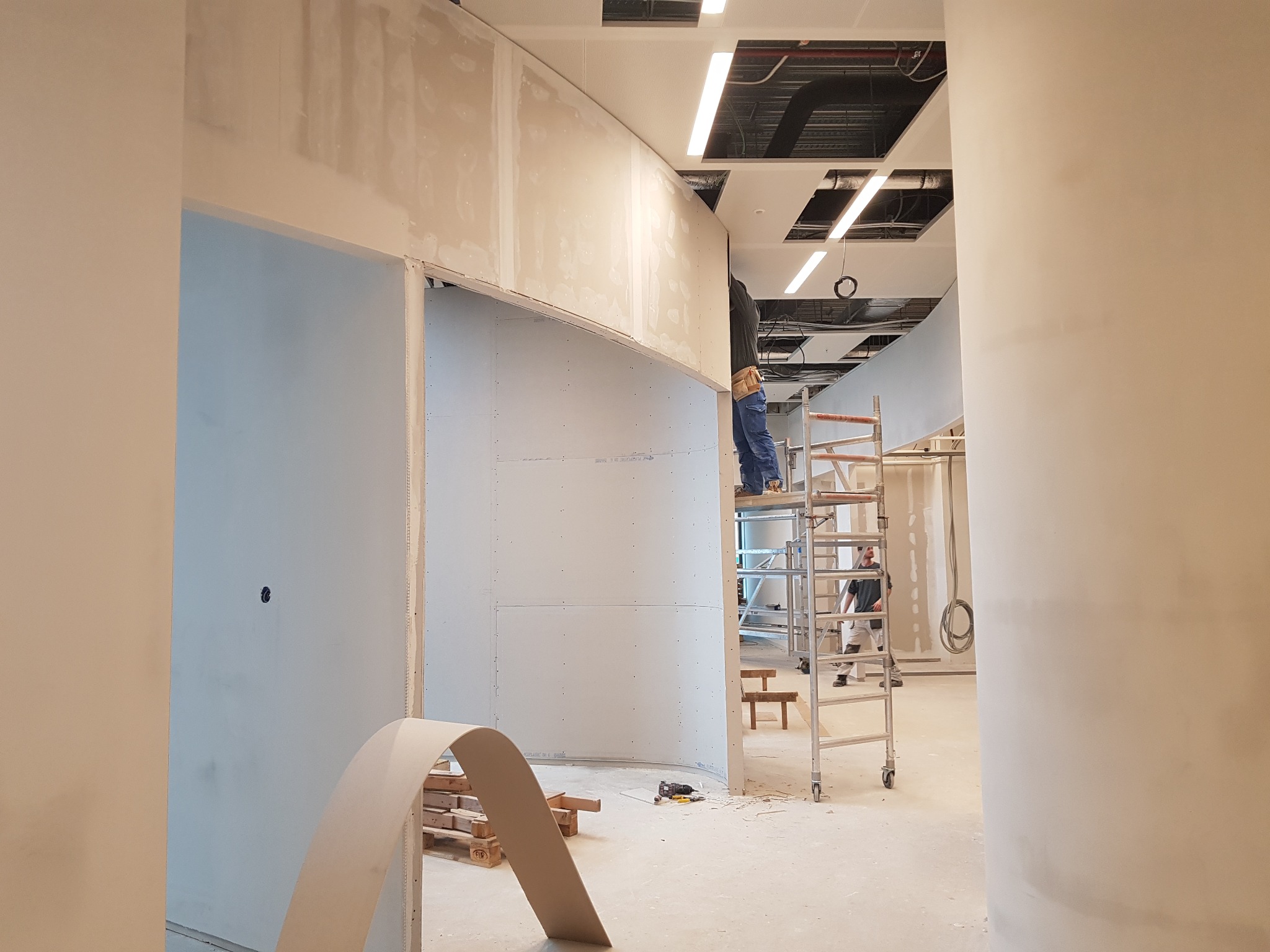 Montaż zaokrąglonej ściany z płyt kartonowo-gipsowych w przestrzeni biurowej, widoczny pracownik na rusztowaniu, narzędzia i fragmenty płyt na podłodze.