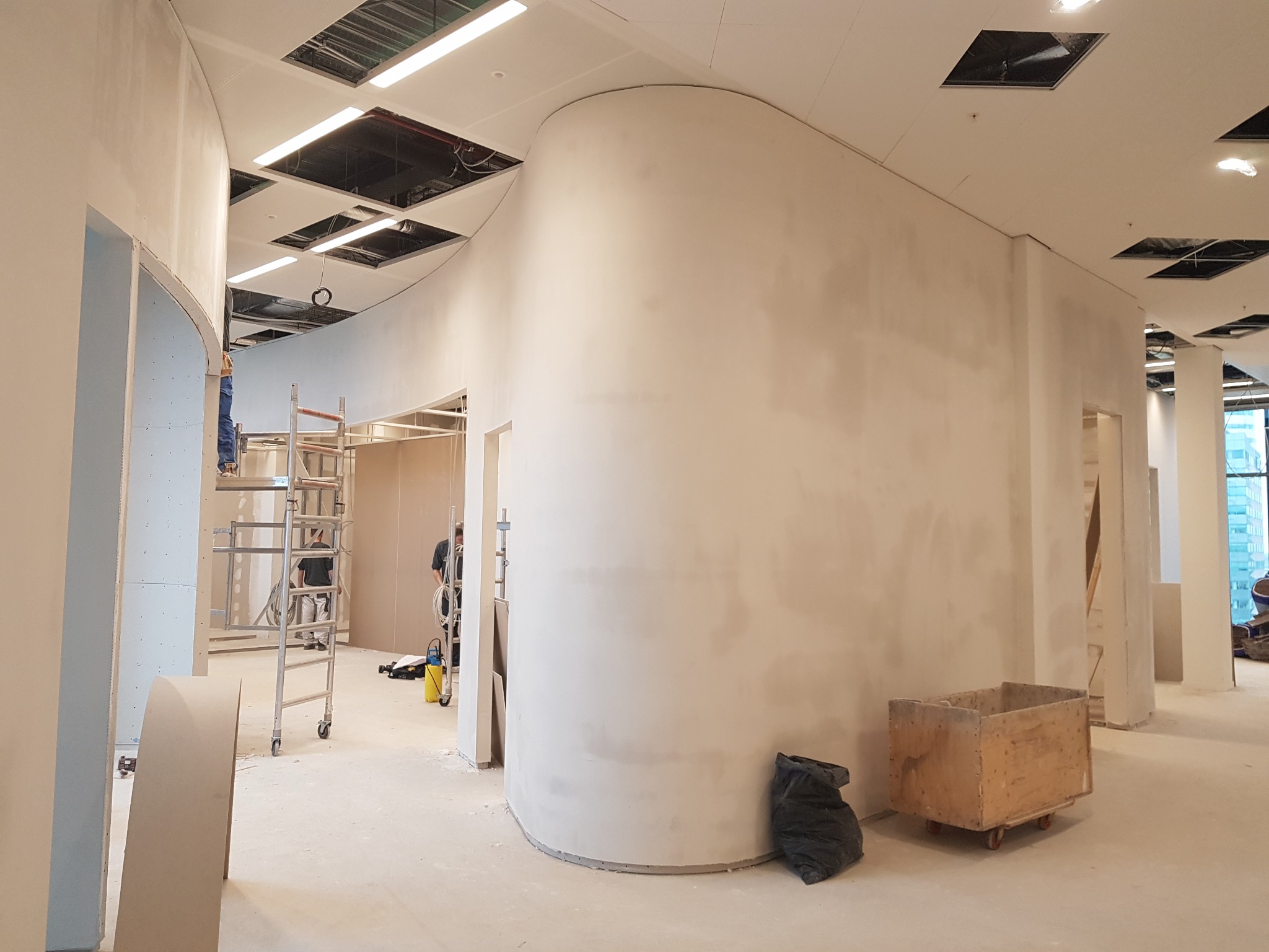Wnętrze biurowe w trakcie wykańczania: zakrzywione ściany z płyt gipsowo-kartonowych, widoczne rusztowania i instalacje pod sufitem podwieszanym.