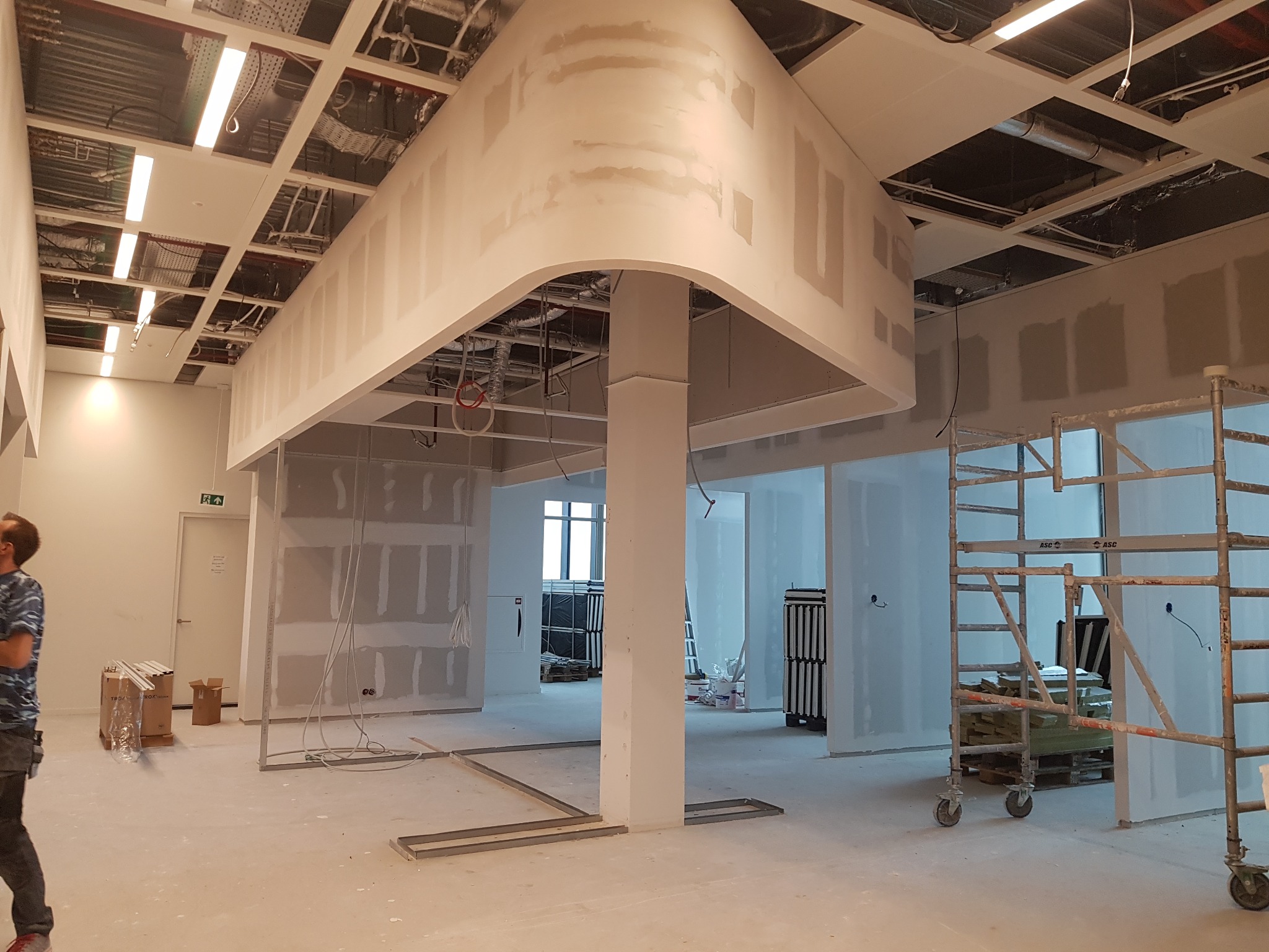 Wnętrze komercyjne w trakcie prac wykończeniowych: widoczna konstrukcja sufitu podwieszanego z elementami karton-gipsu, rusztowanie, przewody elektryczne oraz niedokończone ściany działowe.