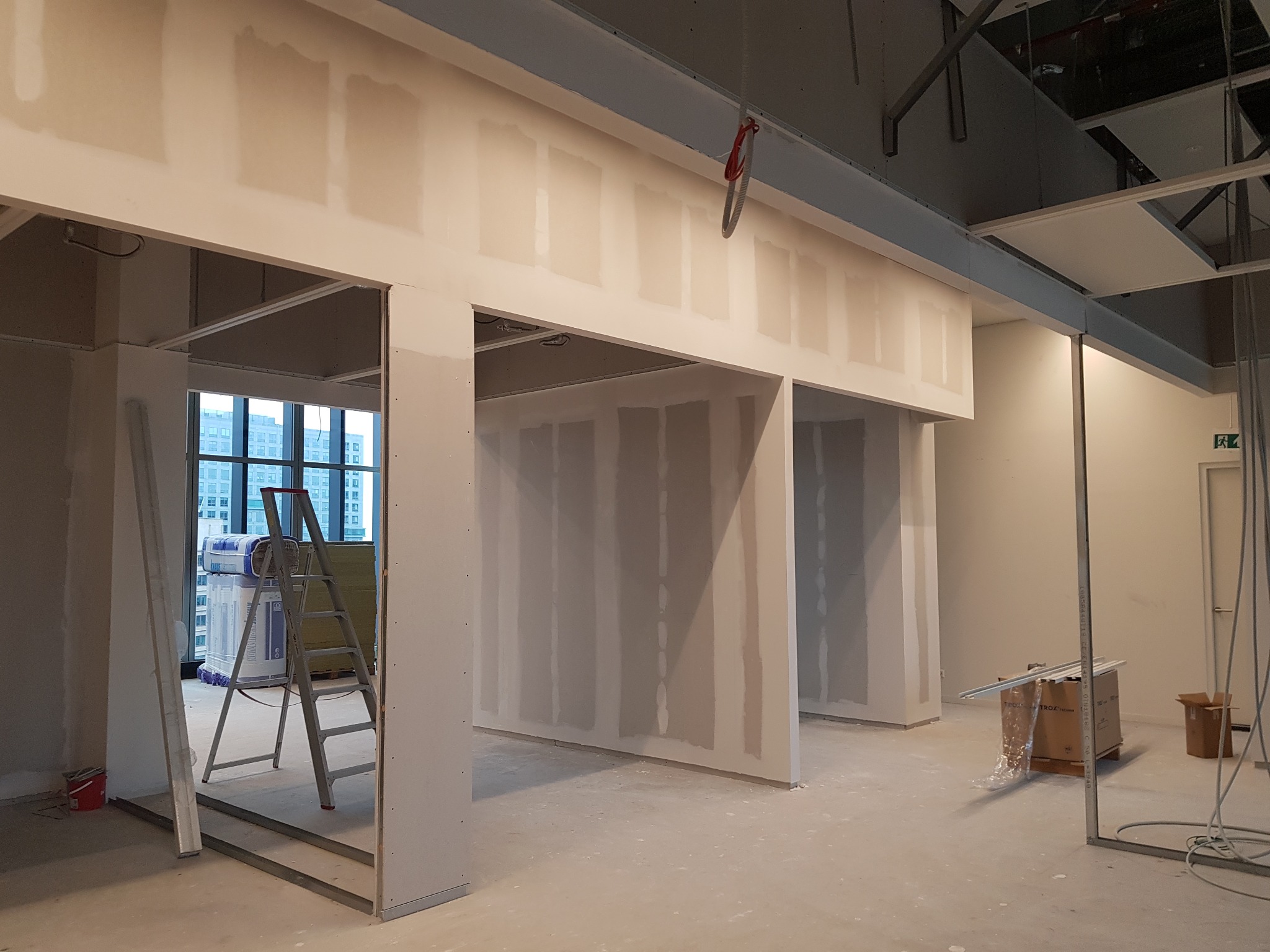 Wnętrze biurowe w trakcie budowy ścian działowych z płyt kartonowo-gipsowych, widoczna drabina oparta o ścianę, paczki z wełną mineralną i metalowe profile.