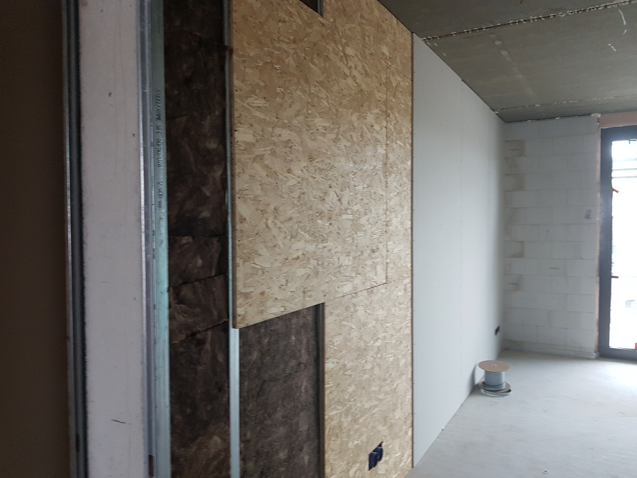 Montaż płyty OSB na ścianie z widoczną izolacją z wełny mineralnej i metalowym stelażem, w tle ściana z bloczków i okno.