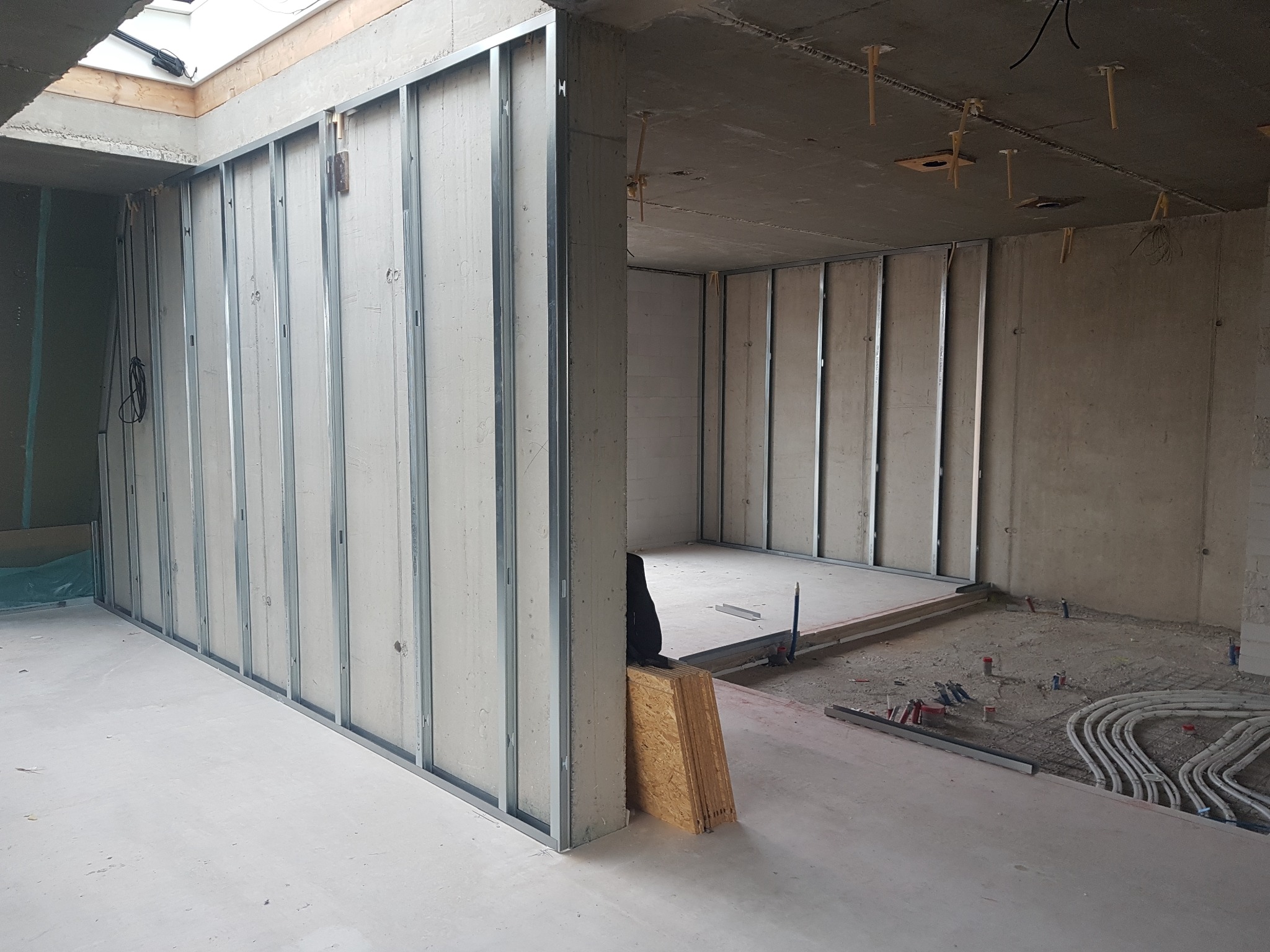 Wnętrze w trakcie prac wykończeniowych: widoczne metalowe profile ścian działowych z płyt kartonowo-gipsowych, instalacje elektryczne w podłodze oraz okno dachowe w surowym stanie.