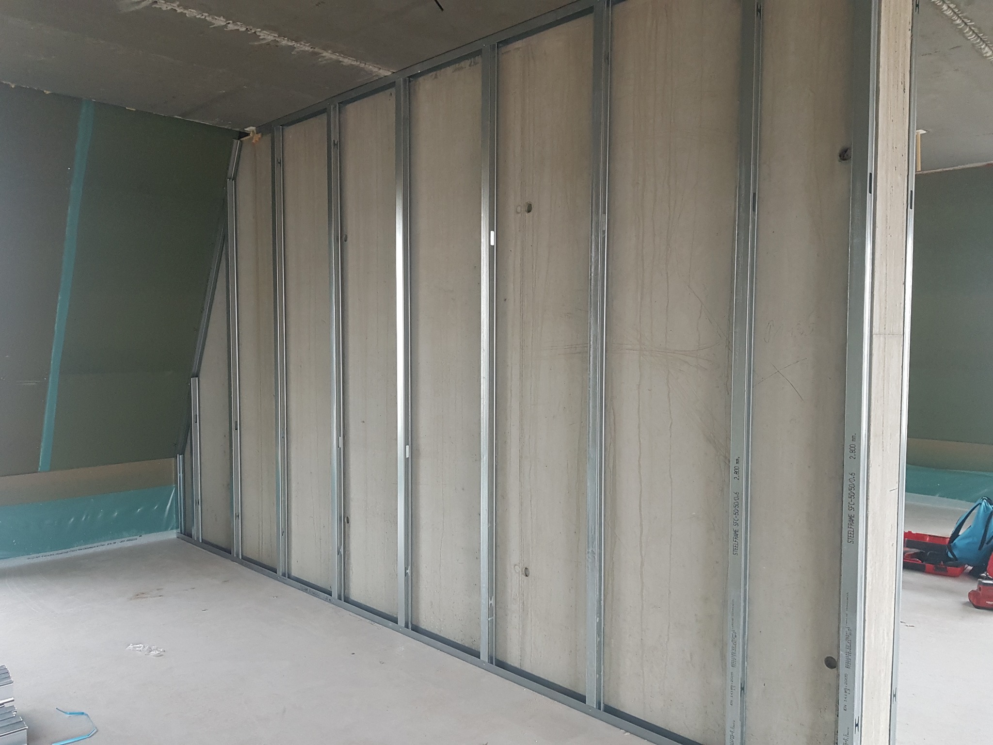 Wnętrze w trakcie prac: metalowy szkielet pod płytę kartonowo-gipsową, częściowo obłożony płytami, widoczny fragment ściany z zielonej płyty gipsowej, surowy betonowy sufit, w tle narzędzia.