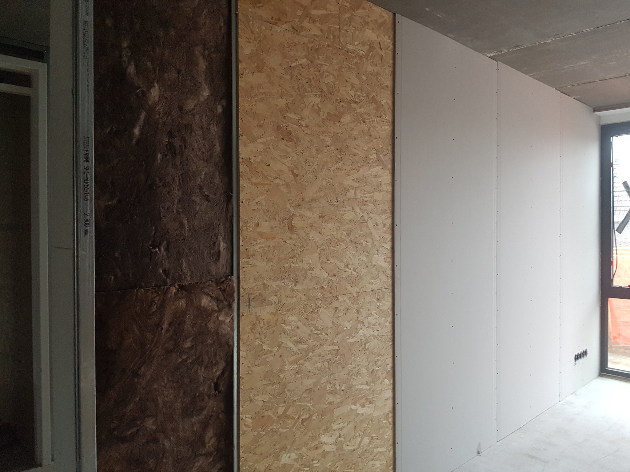 Wnętrze w trakcie prac wykończeniowych: izolacja z wełny mineralnej, płyta OSB i zamontowana płyta gipsowo-kartonowa na metalowym stelażu, widoczne puszki elektryczne.