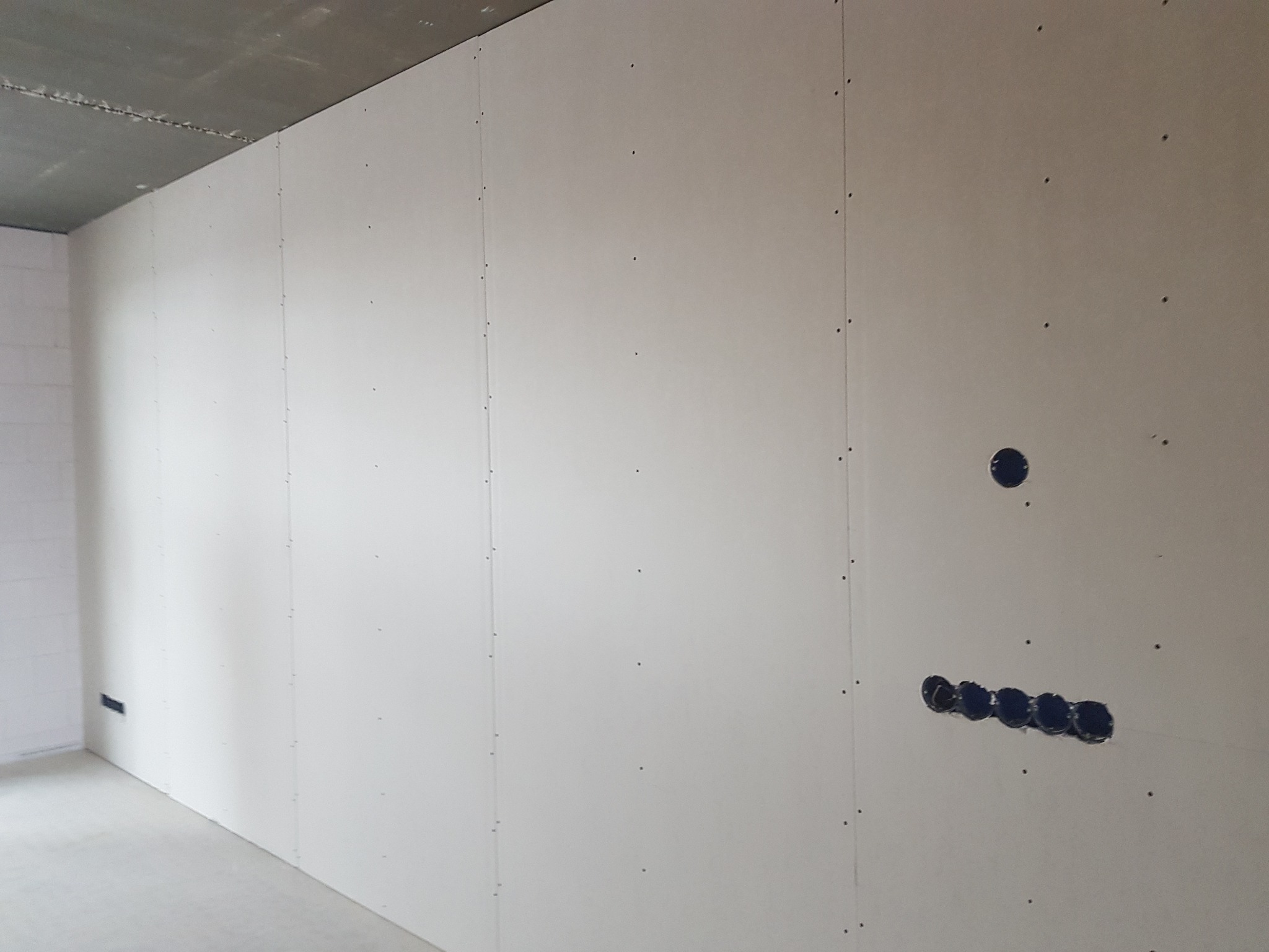 Ściana w surowym stanie z płyt kartonowo-gipsowych, widoczne otwory pod gniazdka elektryczne, szpachlowane łączenia płyt i surowy betonowy sufit.