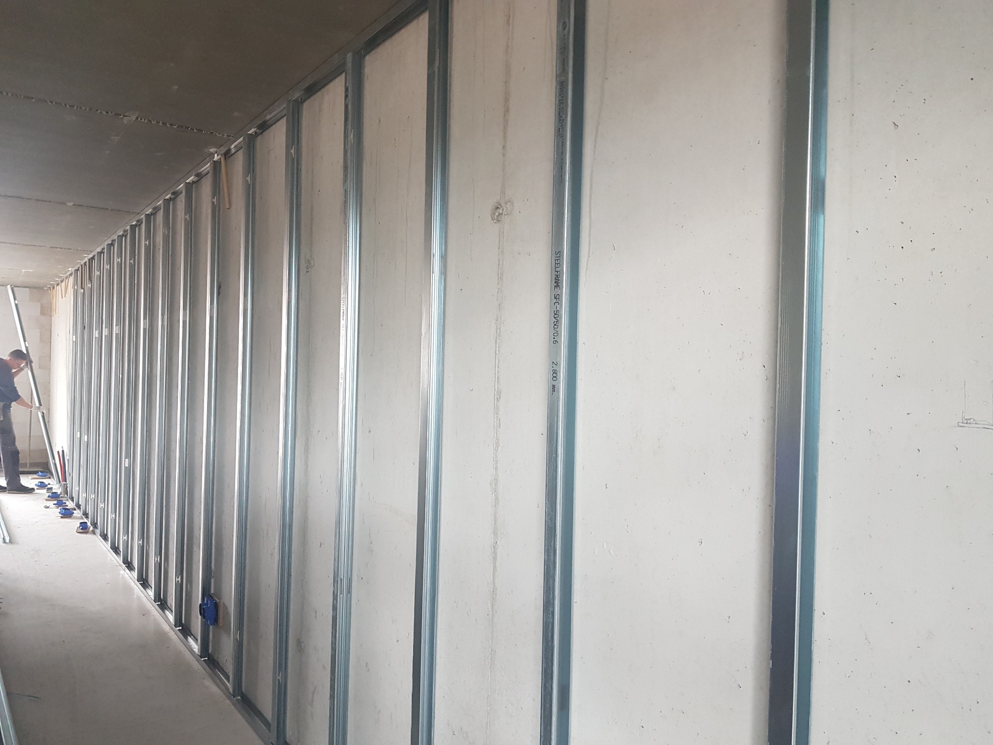 Montaż metalowego stelażu pod płyty gipsowo-kartonowe w surowym wnętrzu, widoczne niebieskie elementy mocujące i pracownik w oddali.