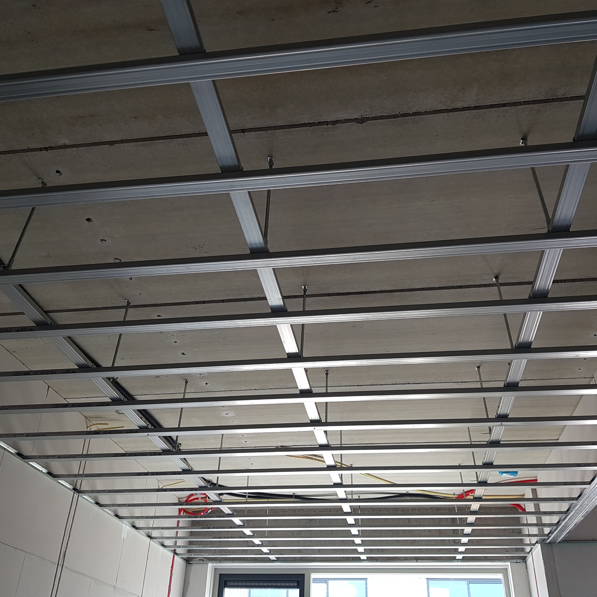 Stelaż sufitu podwieszanego z płyt kartonowo-gipsowych, widoczne profile stalowe, betonowy strop powyżej, instalacje i fragment okna w tle.