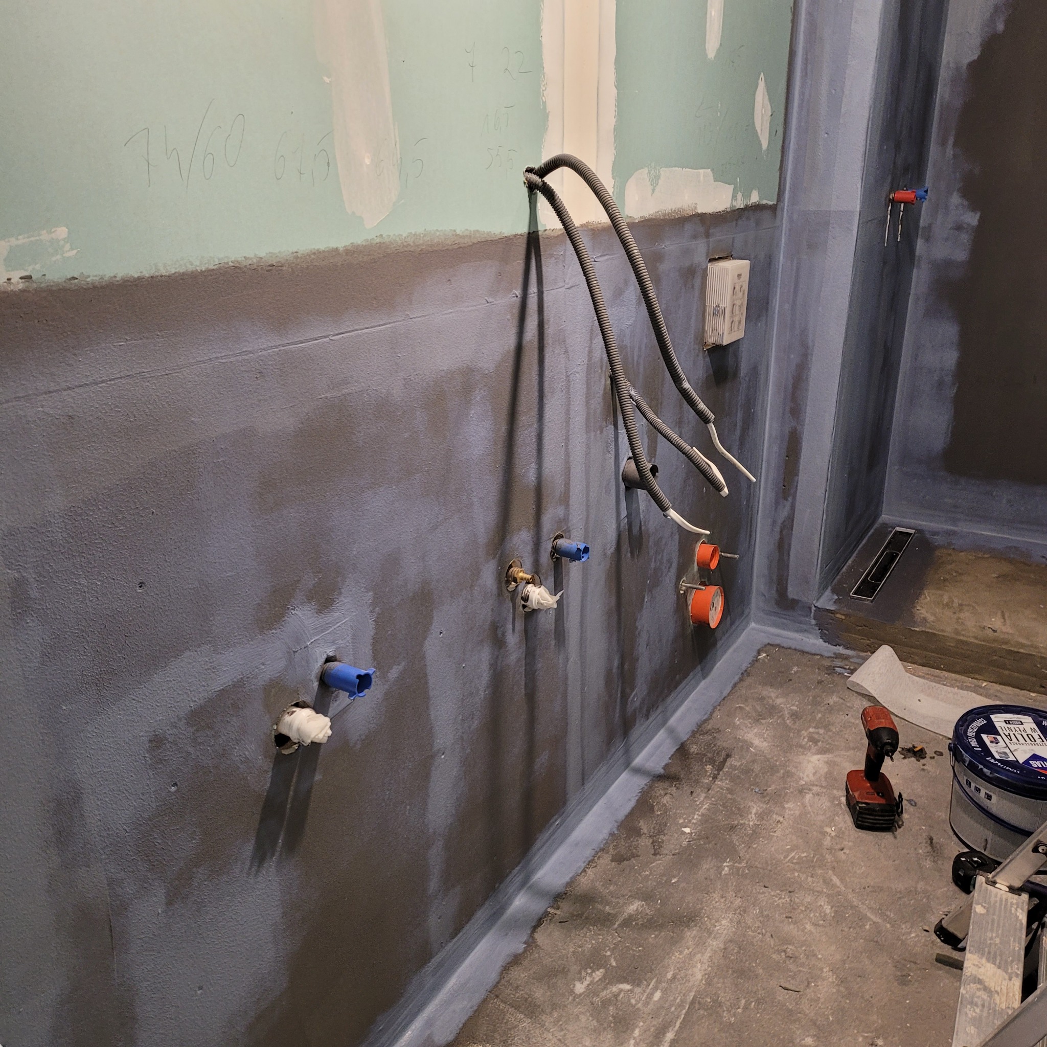 Przygotowana do wykończenia łazienka z nałożoną hydroizolacją, widoczne wystające rury, instalacje, odpływ liniowy, narzędzia, surowe ściany z płyt kartonowo-gipsowych z naniesionymi notatkami.