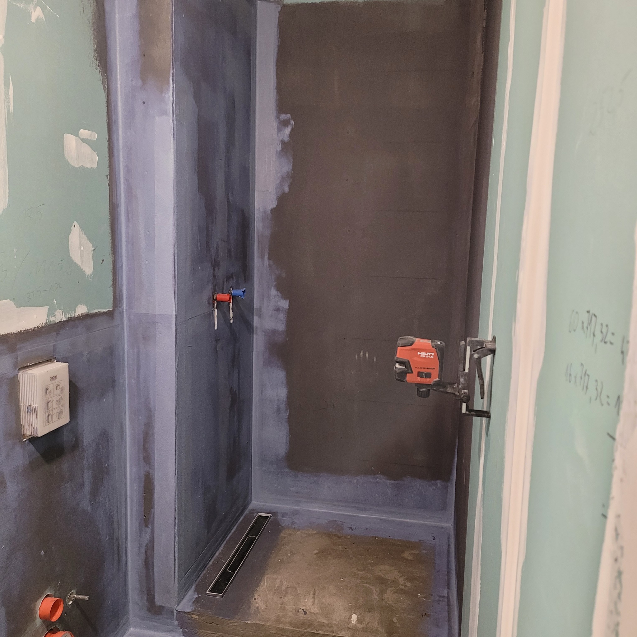 Przygotowanie do hydroizolacji w narożnej kabinie prysznicowej z odpływem liniowym, widoczne pionowe rury instalacyjne, laser krzyżowy Hilti PM 2-LG zamocowany na ścianie z płyt kartonowo-gipsowych...