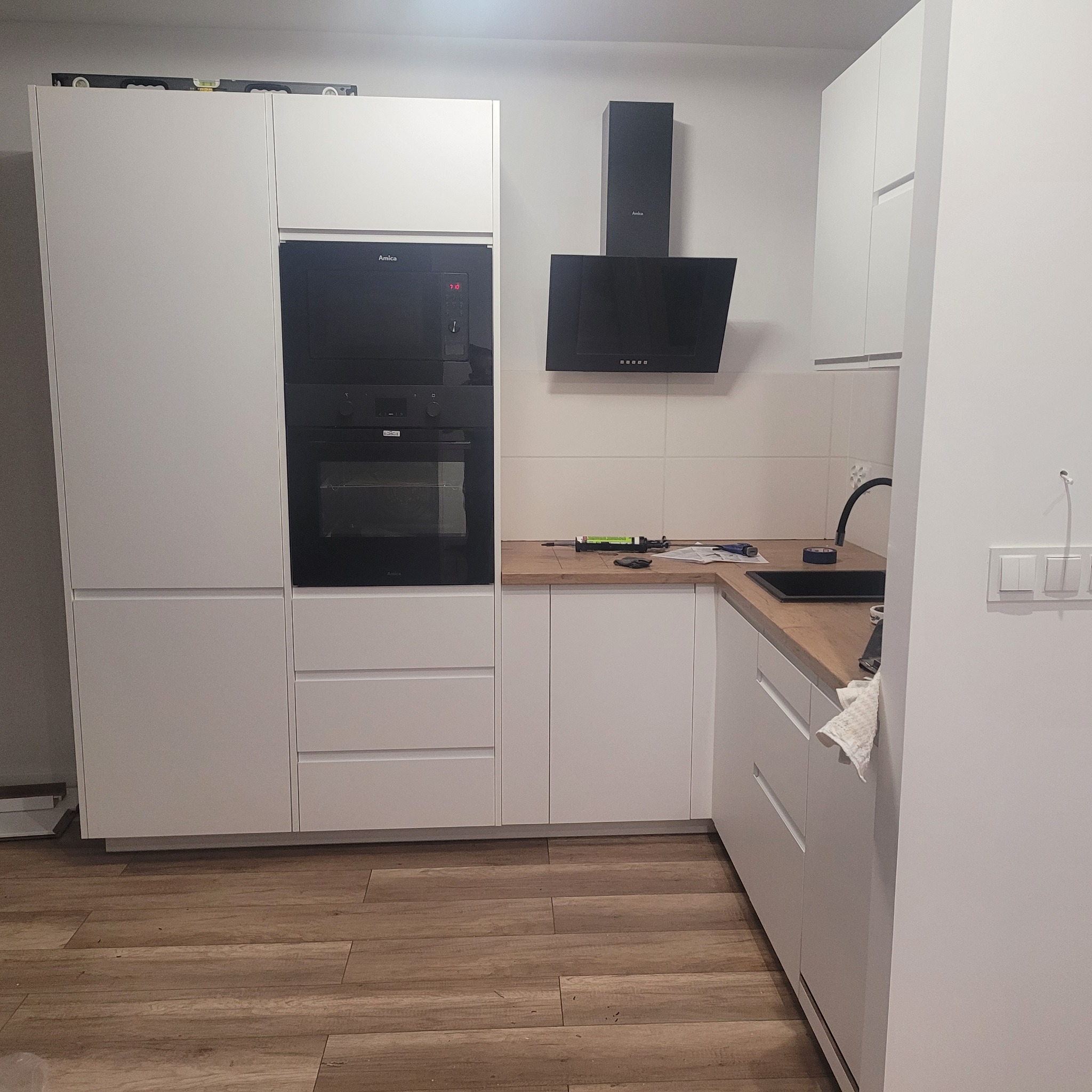 Nowoczesna, biała zabudowa kuchenna z wbudowanym piekarnikiem i płytą grzewczą, czarnym okapem oraz drewnianym blatem, na którym leżą narzędzia i akcesoria montażowe, co sugeruje trwający montaż.