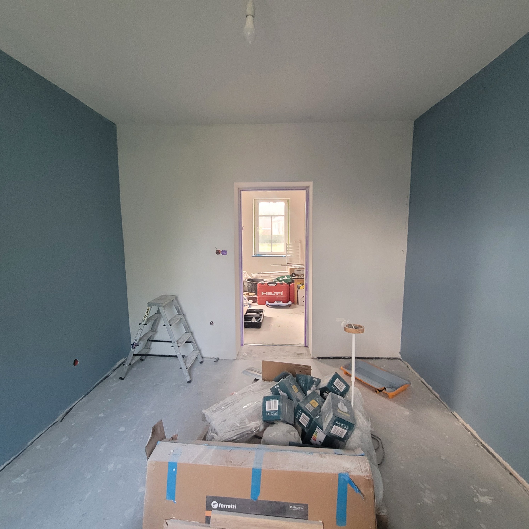 Pokój po malowaniu ścian na biało i niebiesko. Widoczna drabina, kartony z panelami i narzędzia malarskie. Przez drzwi widać dalszą część remontowanego mieszkania.