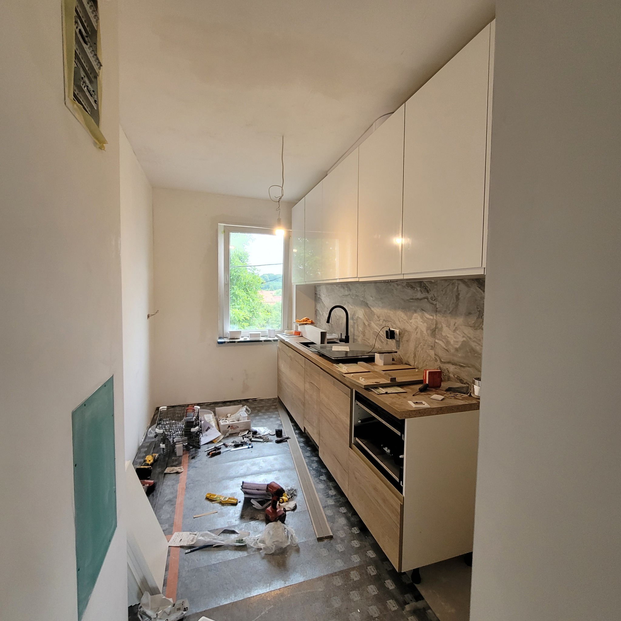 Wnętrze kuchni w trakcie montażu: białe szafki wiszące, drewnopodobne dolne, okno z widokiem na zieleń. Na podłodze narzędzia i materiały budowlane, ściany przygotowane do malowania.