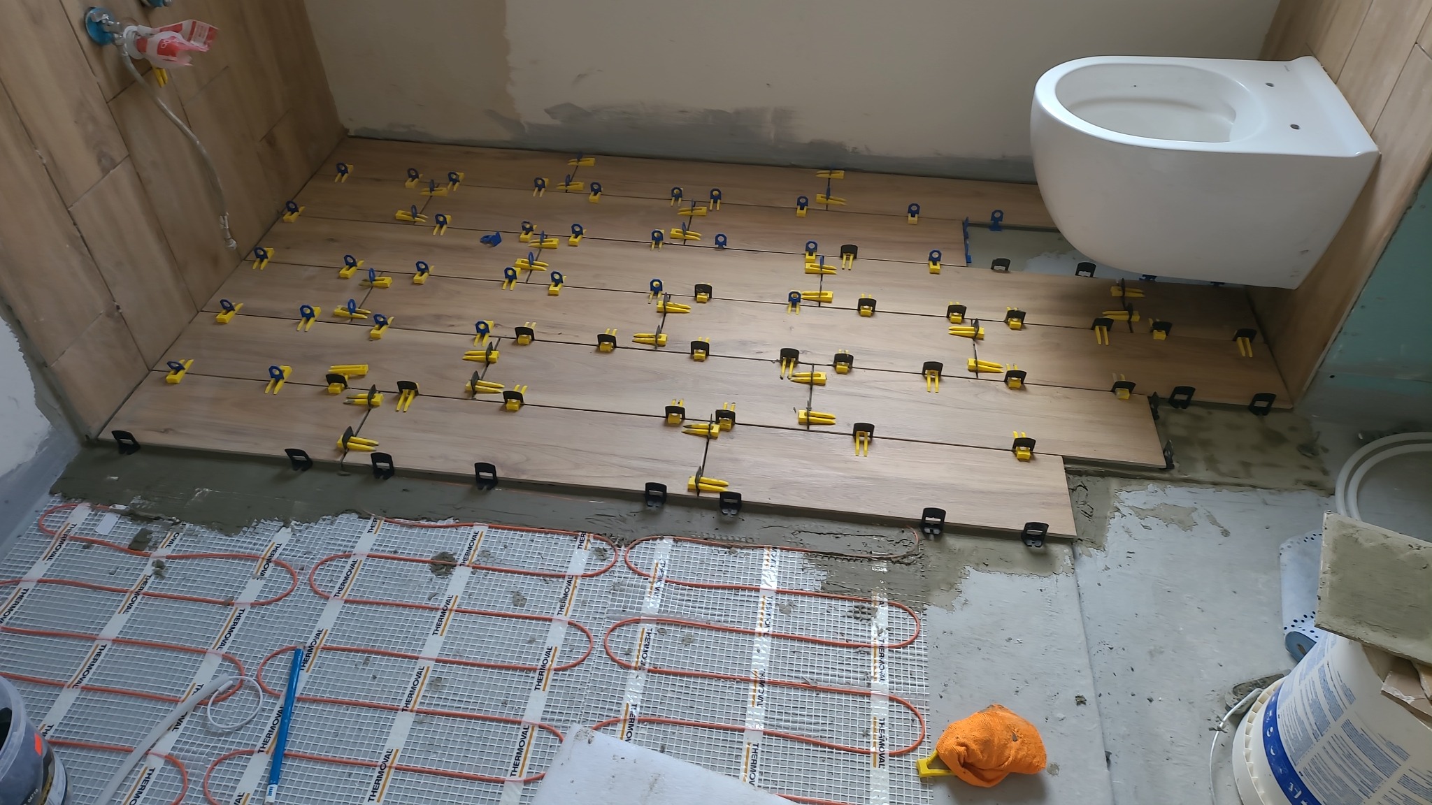 Układanie płytek imitujących drewno w łazience z zamontowaną toaletą. Widoczne żółte klipsy poziomujące i mata grzewcza pod płytkami.