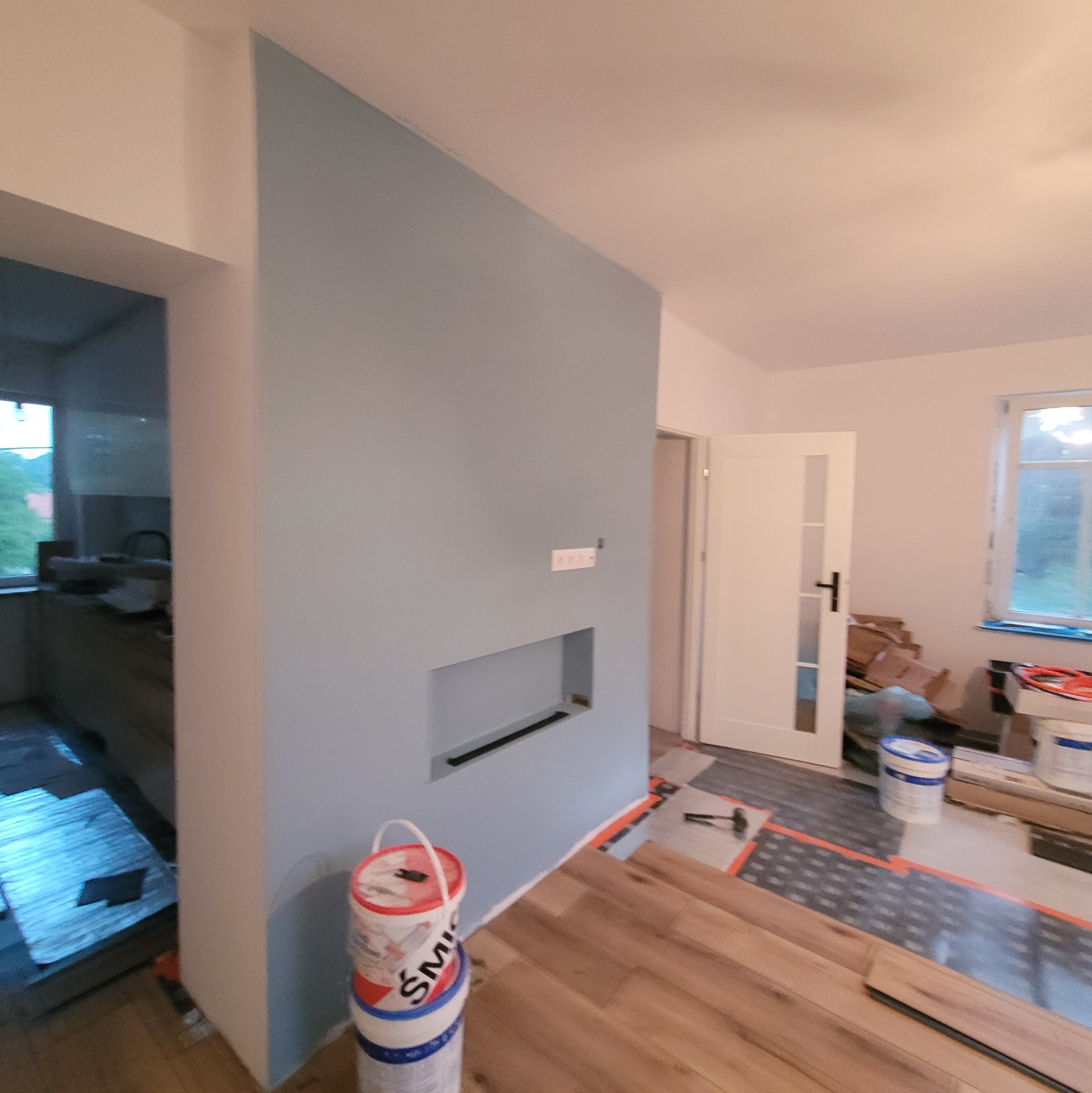 Ściana pomalowana na błękitno z wnęką, częściowo ułożone panele podłogowe, wiadro z farbą. Widoczny fragment kuchni i drzwi. Wnętrze w trakcie wykańczania.