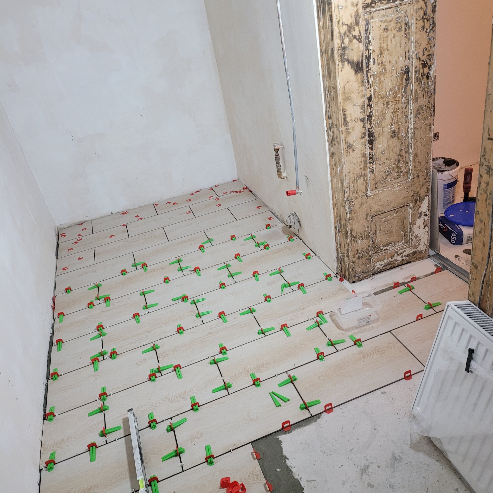 Układanie jasnych płytek podłogowych z użyciem zielonych i czerwonych klipsów poziomujących w trakcie remontu. Widoczna stara, obdrapana futryna drzwi.