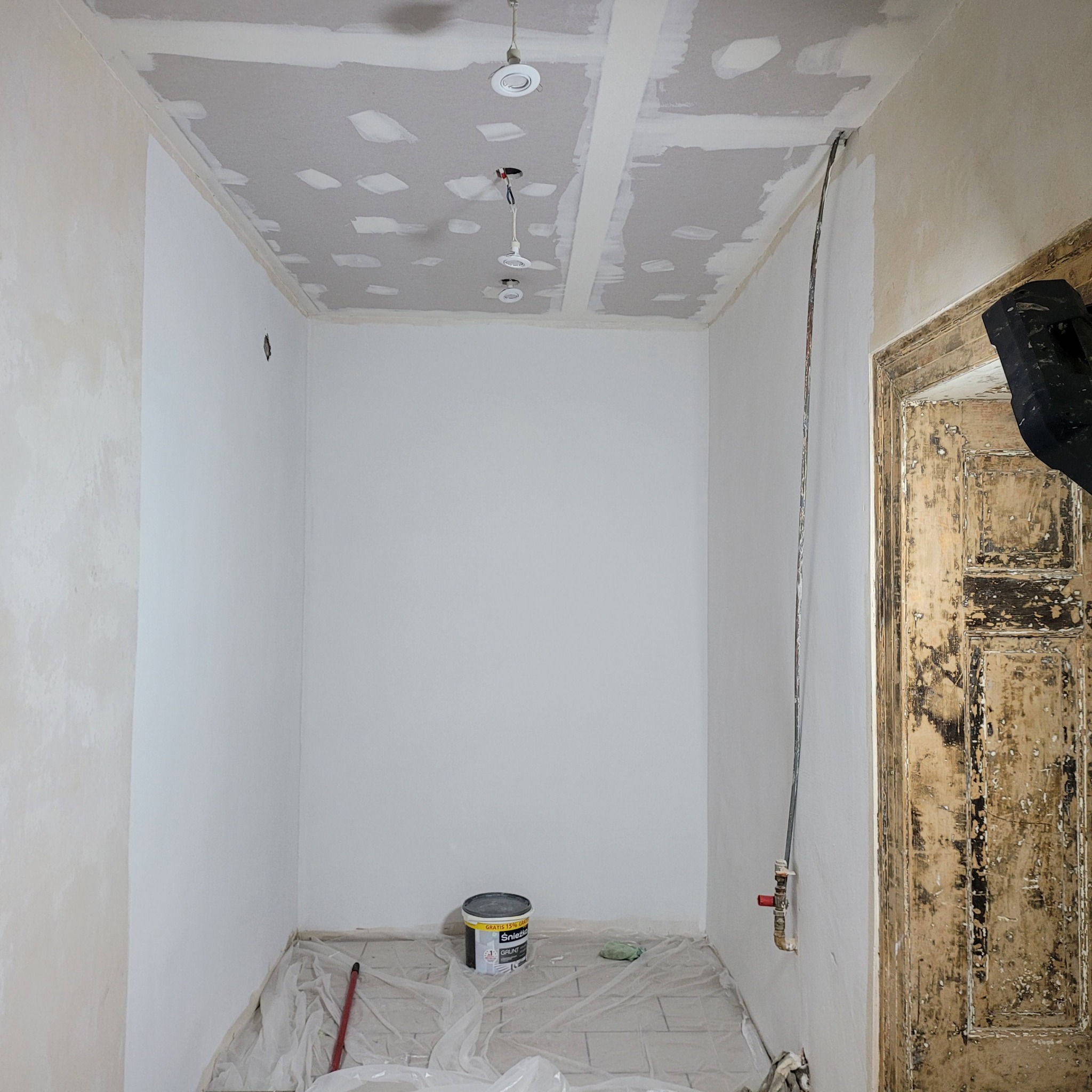 Wnętrze w trakcie prac wykończeniowych: ściany pomalowane na biało, sufit z płyt karton-gips, widoczne punkty oświetleniowe i wiadro z farbą. Stara, odrapana futryna.