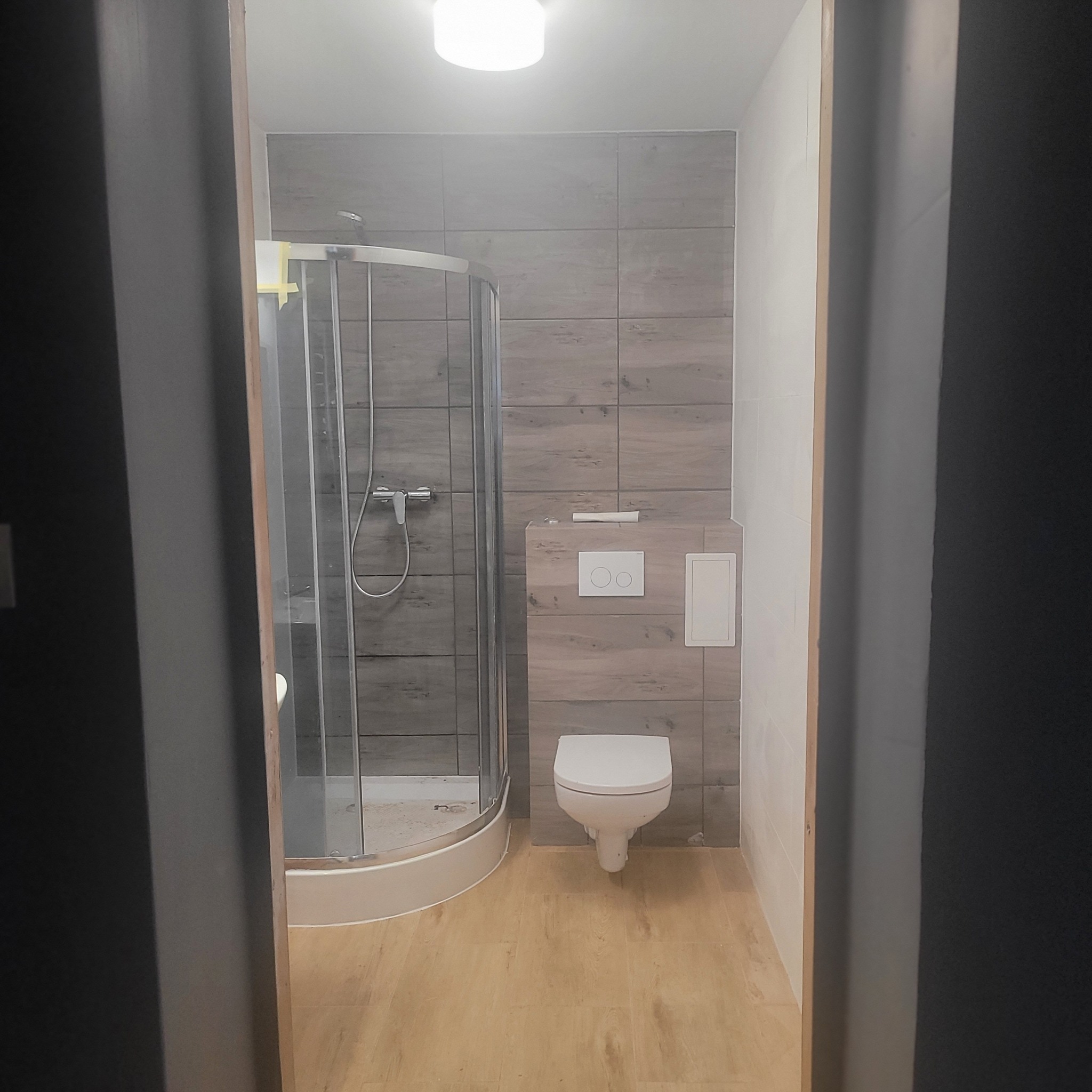 Wykończona łazienka z kabiną prysznicową, toaletą i płytkami imitującymi drewno na ścianach. Widok zza framugi drzwi.