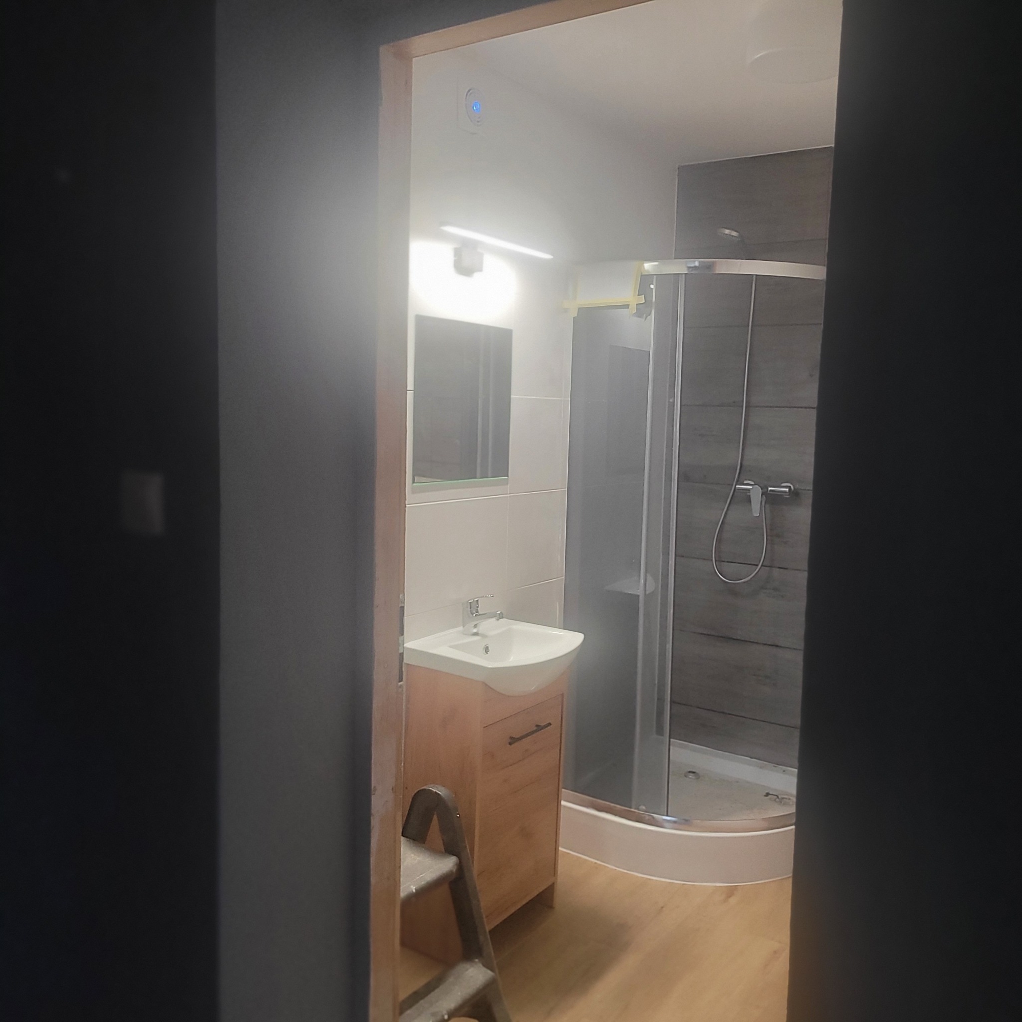 Nowoczesna łazienka po remoncie: kabina prysznicowa z szarymi płytkami imitującymi drewno, umywalka z szafką w odcieniach drewna, widok zza futryny.