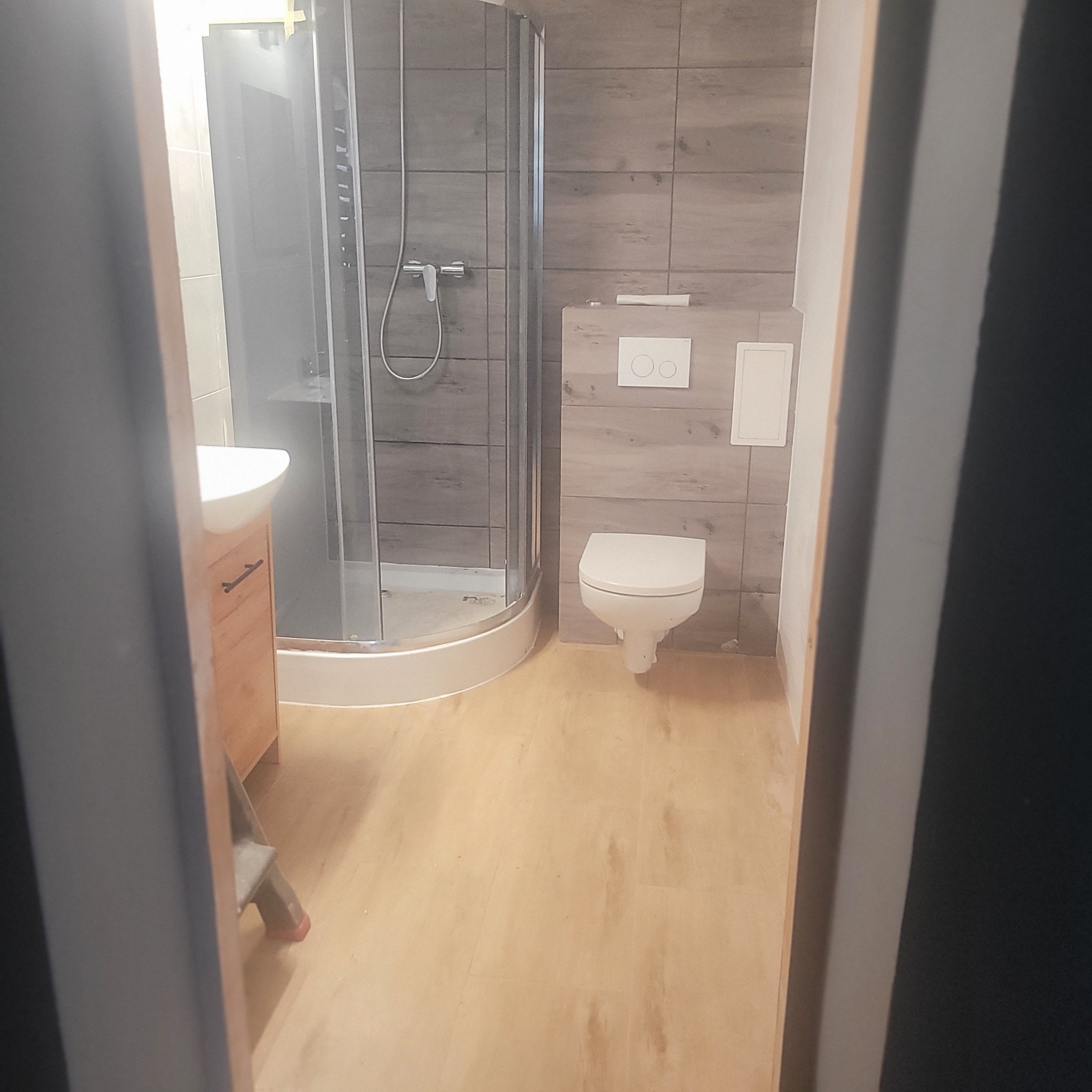 Wykończona łazienka z kabiną prysznicową, toaletą i umywalką z szafką. Płytki imitujące drewno na ścianach i podłodze. Widok zza framugi drzwi.
