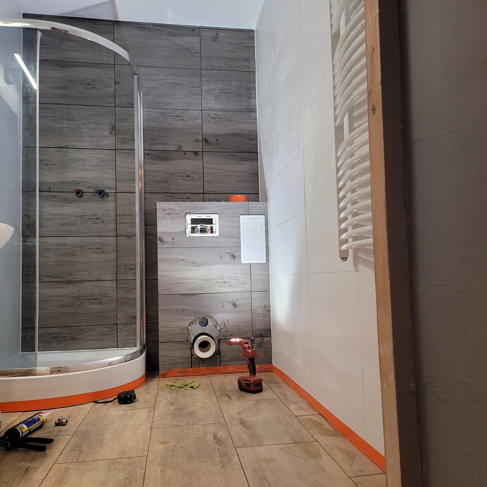 Montaż podtynkowej toalety w łazience z szarymi płytkami imitującymi drewno i białą glazurą. Widoczna kabina prysznicowa, narzędzia i grzejnik drabinkowy.