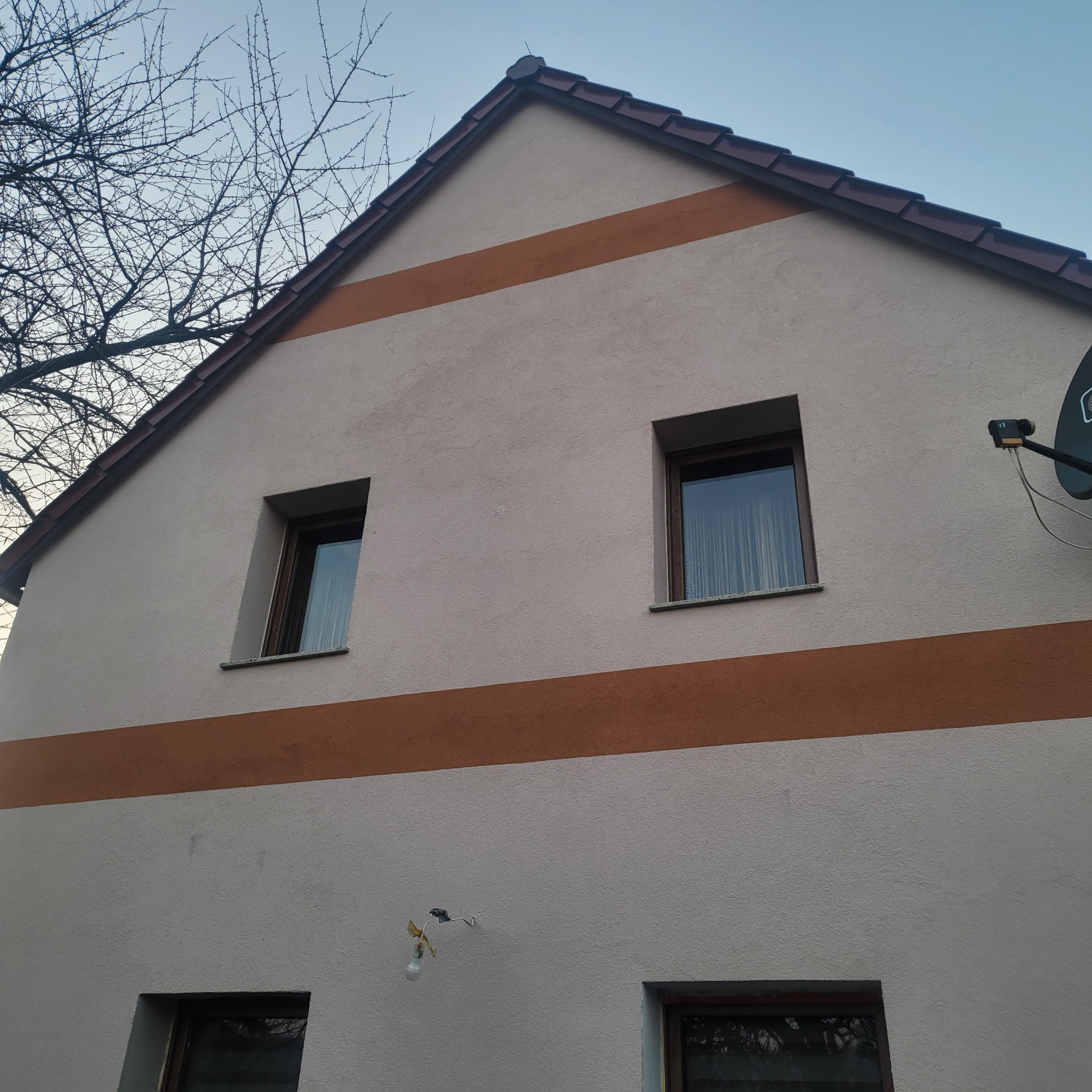Elewacja domu jednorodzinnego z dwoma pasami w kolorze brązowym. Widoczne okna i antena satelitarna. Na pierwszym planie gałęzie drzewa.