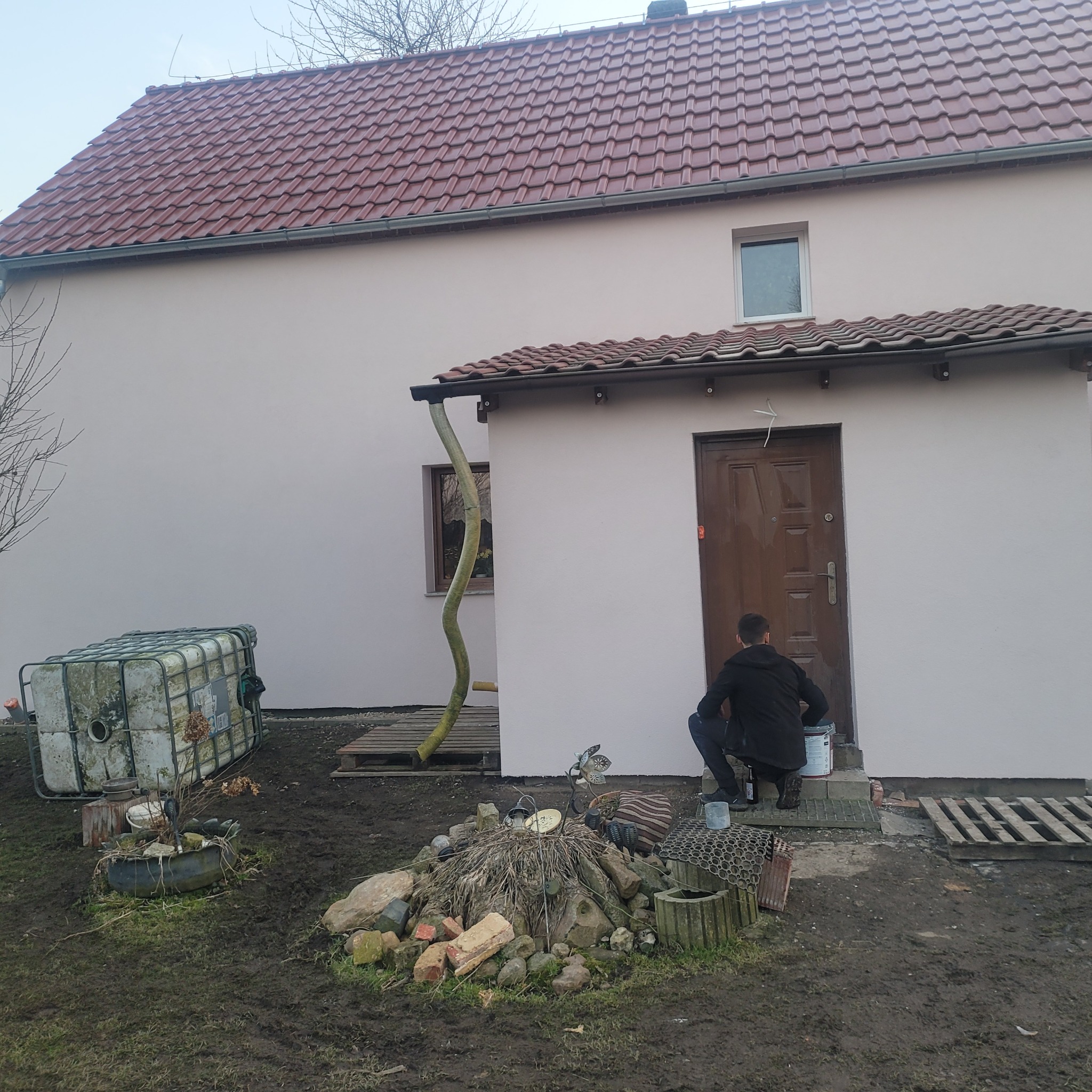 Mężczyzna maluje elewację domu na jasny kolor. Widoczna puszka z farbą, fragment dachu z czerwoną dachówką i otoczenie budynku.