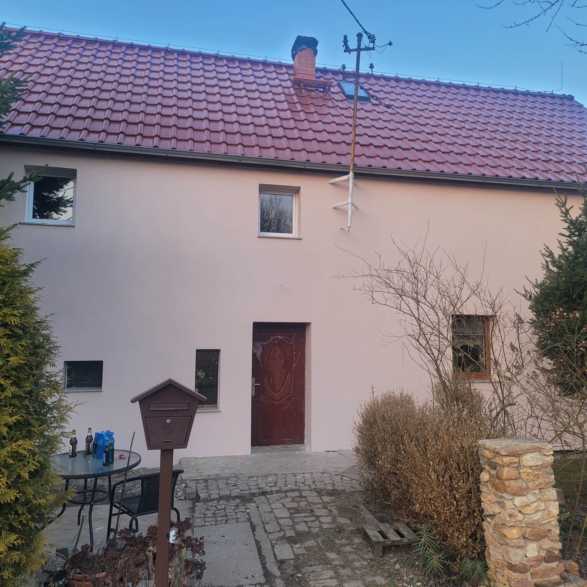 Odnowiony dom jednorodzinny z czerwoną dachówką i świeżo pomalowaną elewacją w odcieniach beżu. Widoczny fragment ogrodu z meblami i skrzynką pocztową.